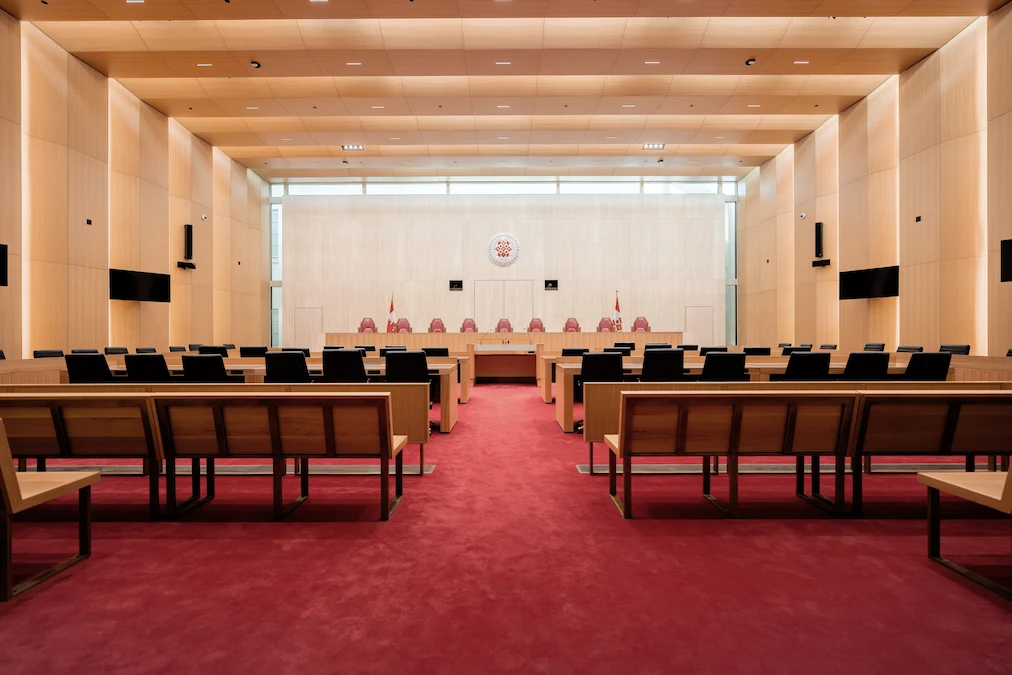 The Supreme Court of Canada courtroom at the West Memorial Building. The ceiling and walls feature wood arches separated by skylights and the carpet is red. The judges’ bench sits at the front of the room.